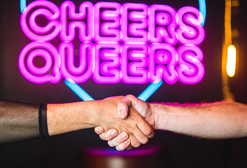Zwei Personen reichen sich die Haende vor dem SchwuZ Neon Leuchtzeichen mit der Aufschrift Cheers Queers